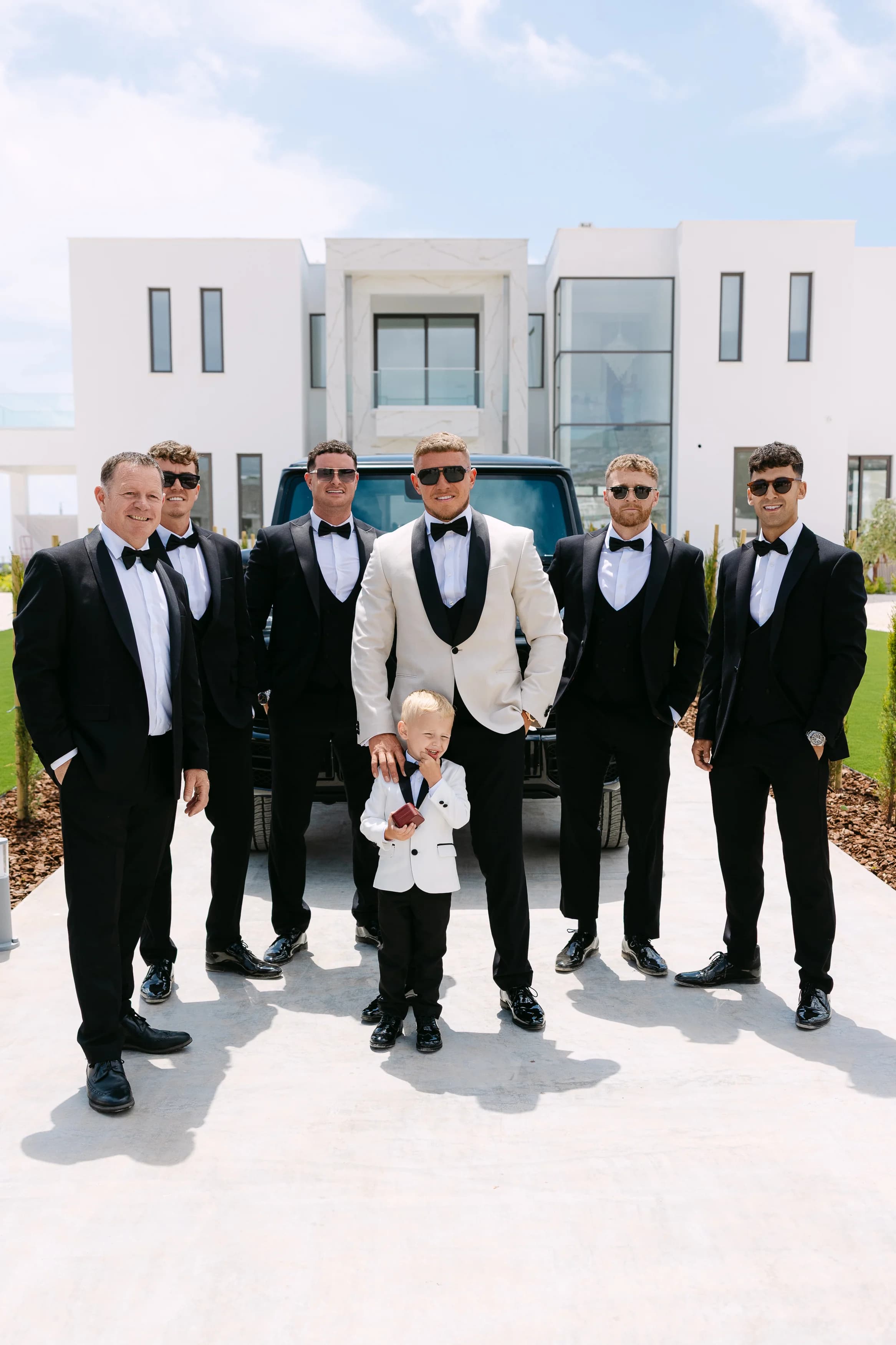 Groom, groomsmen, and young ring bearer photographed in front of the venue with a black Mercedes G-Wagon.