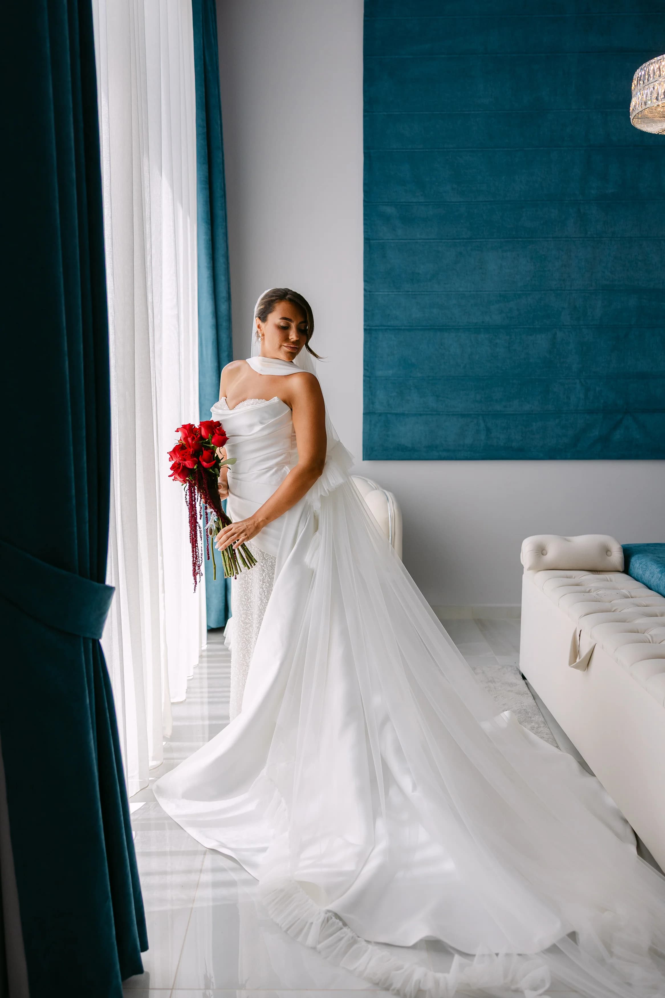 Bride in her wedding gown holding a red bouquet beside the window in the bridal suite at OZEA.