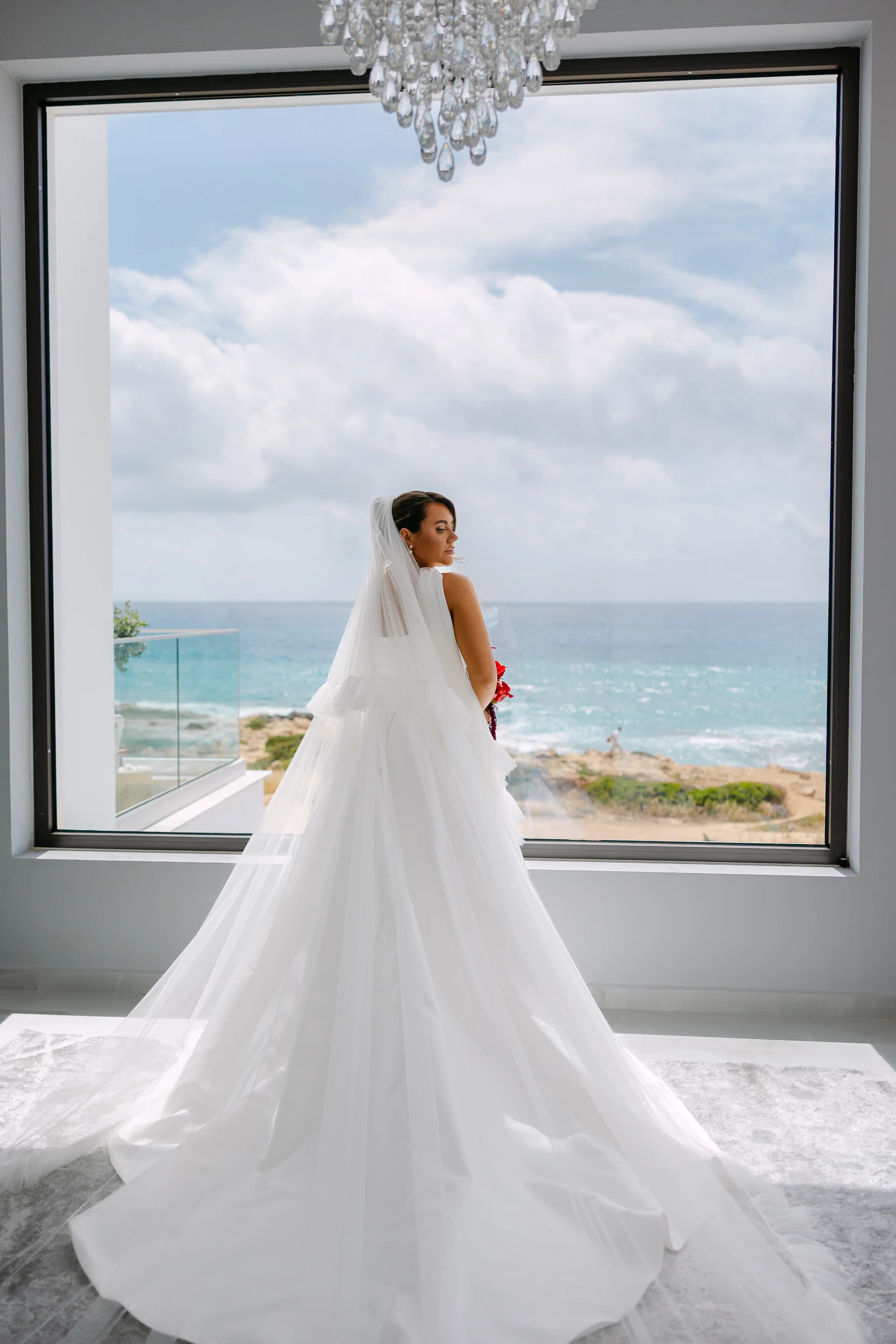 Bride beneath a crystal chandelier facing a panoramic window with the Paphos coastline beyond.