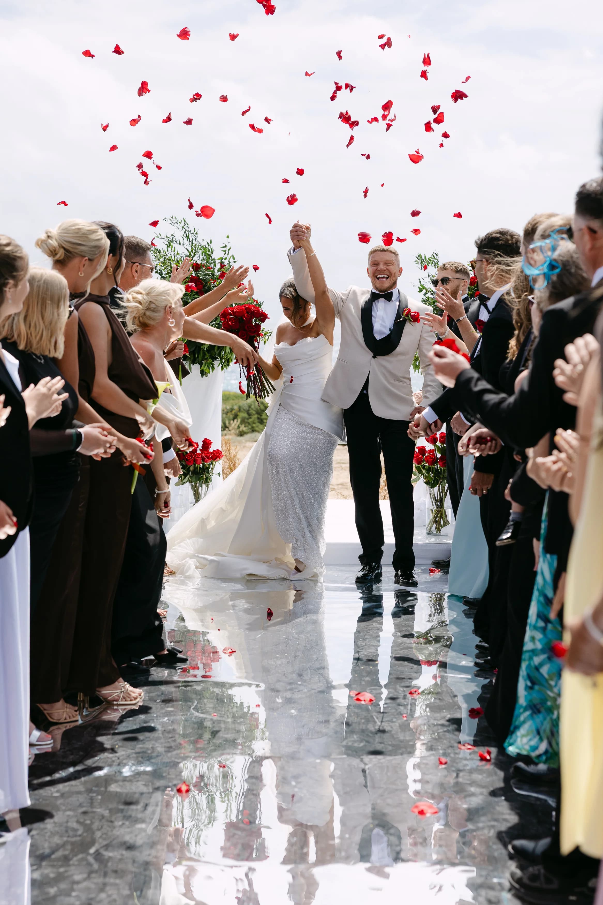 Newlywed couple raising joined hands as red petal confetti falls during their ceremony exit at OZEA.