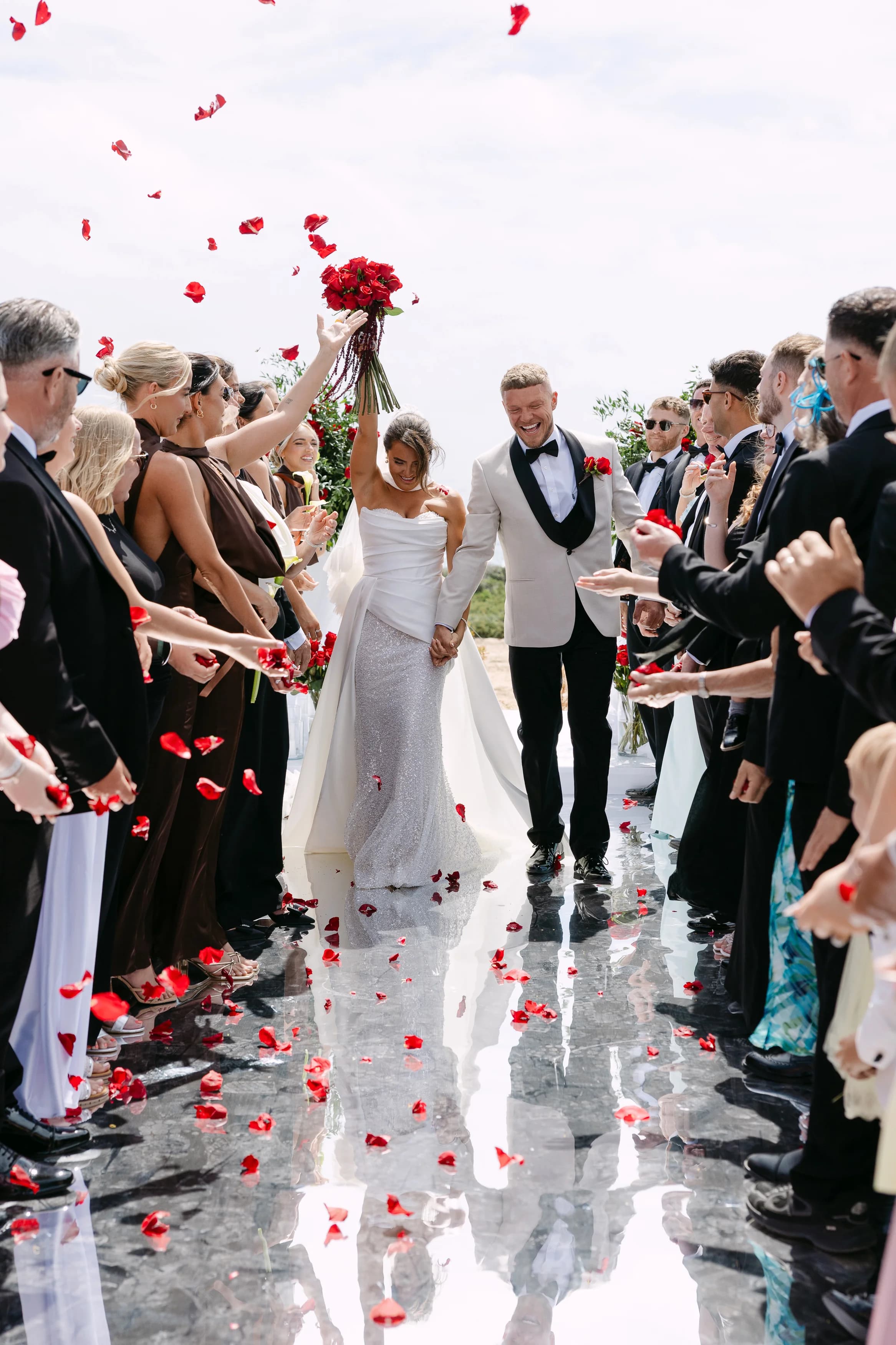 Bride lifting her red rose bouquet during the confetti exit at OZEA, with guests cheering on either side.