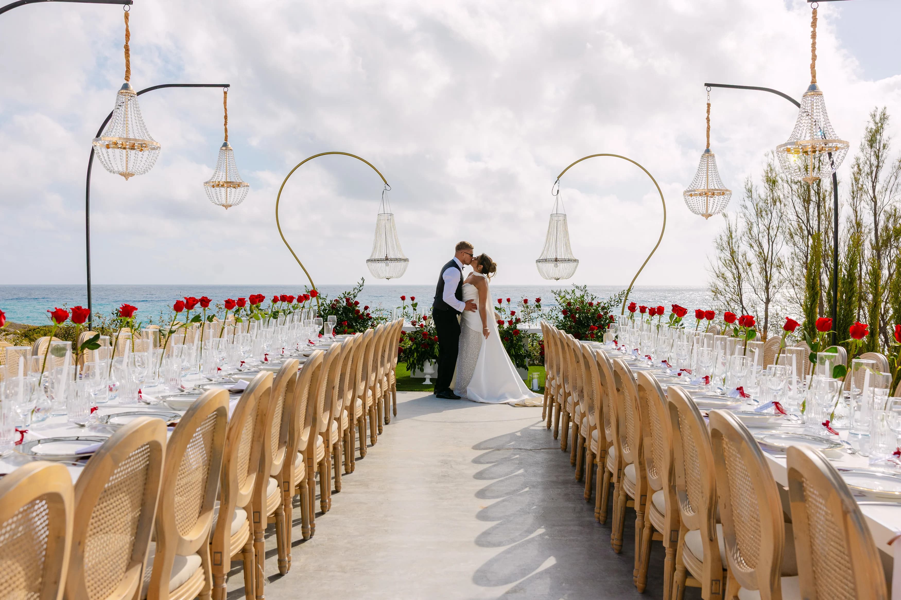 Bride and groom kiss between the long banquet tables at Ozea with chandeliers above and the sea behind.