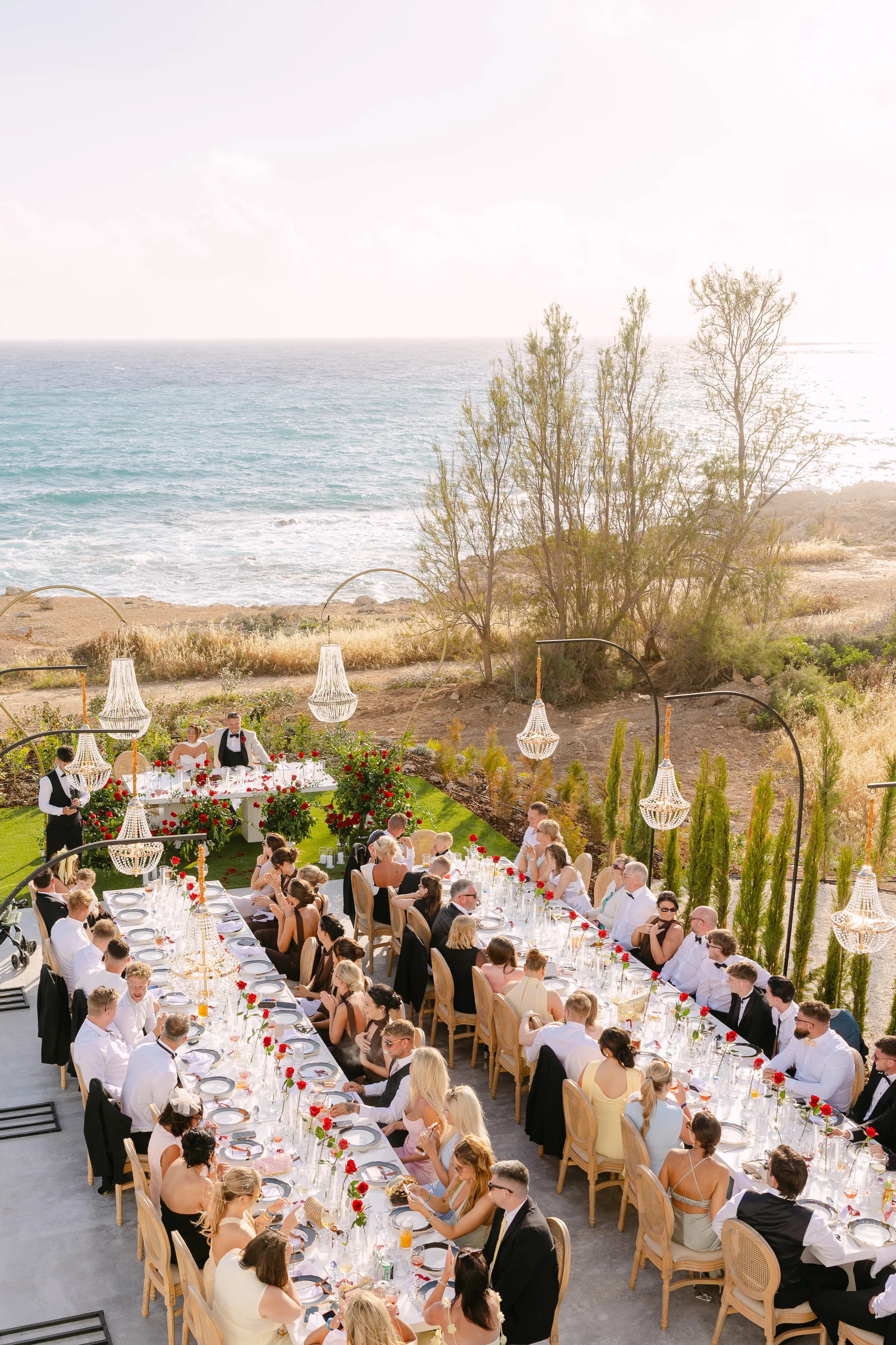 Elevated golden-hour view of a wedding banquet at OZEA with guests seated, chandeliers lit, and the Mediterranean glowing behind.