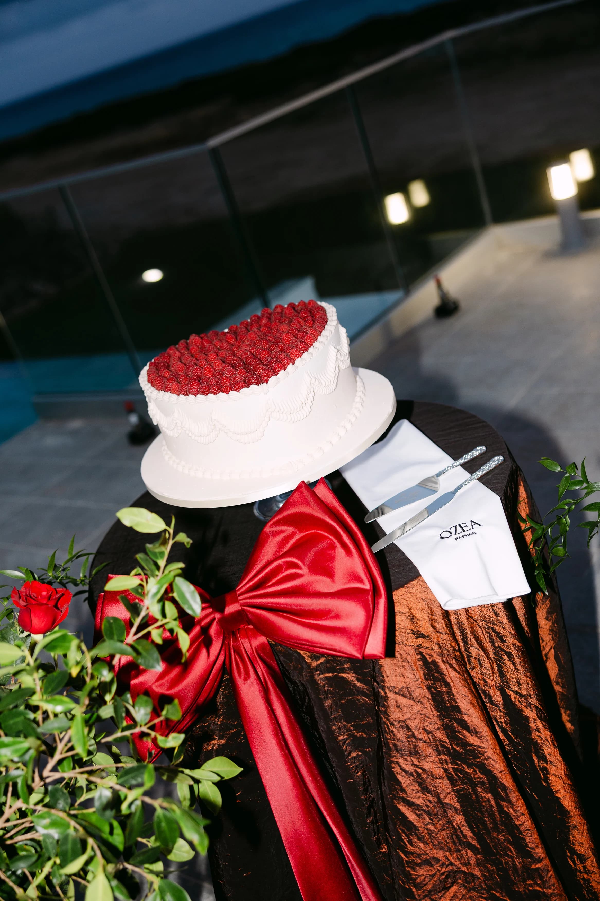 Wedding cake topped with red roses on a black silk-draped table with a wide red bow at the front.