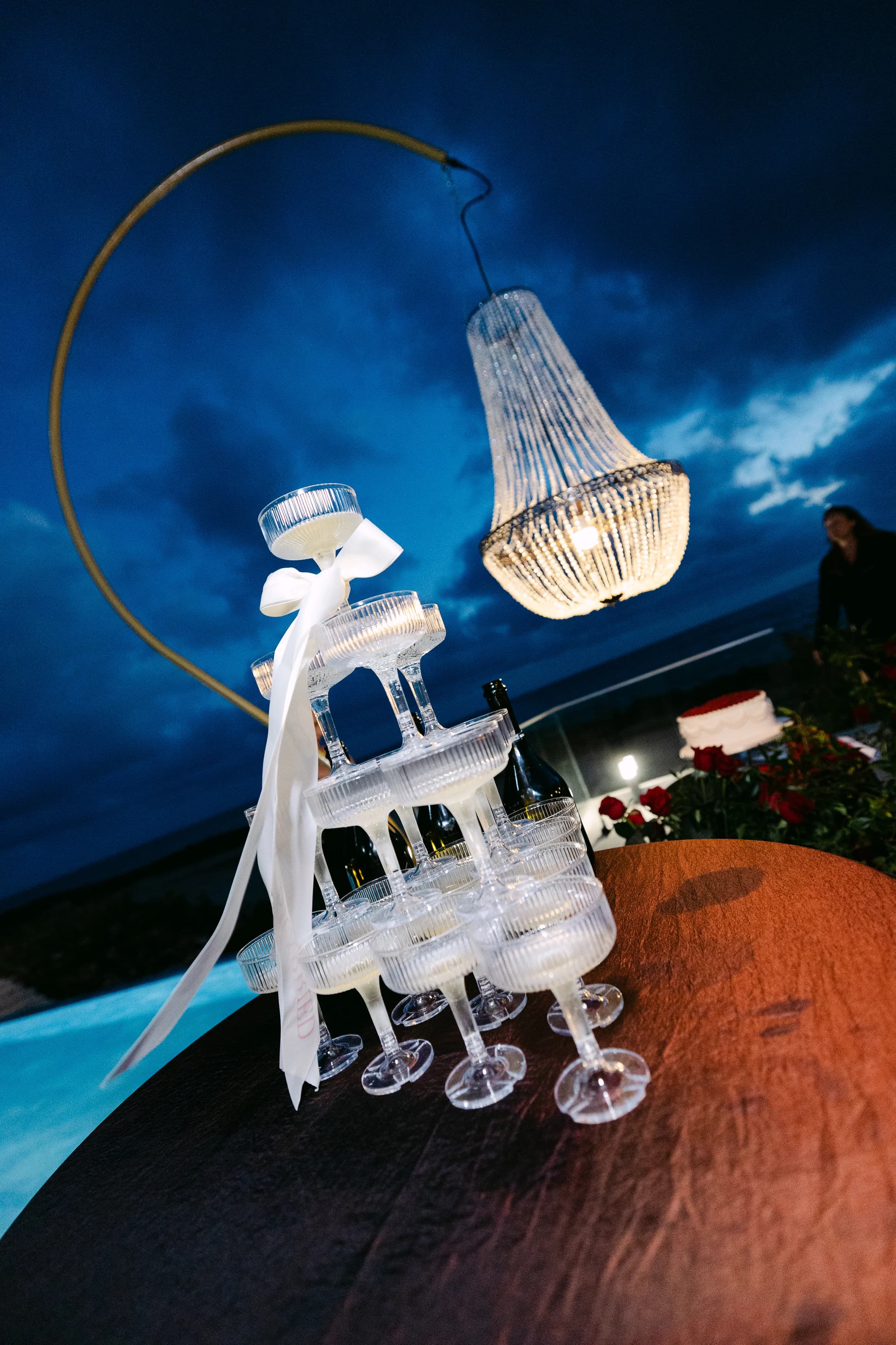 Champagne coupe tower at OZEA photographed at dusk beneath a crystal chandelier, with the sea behind.