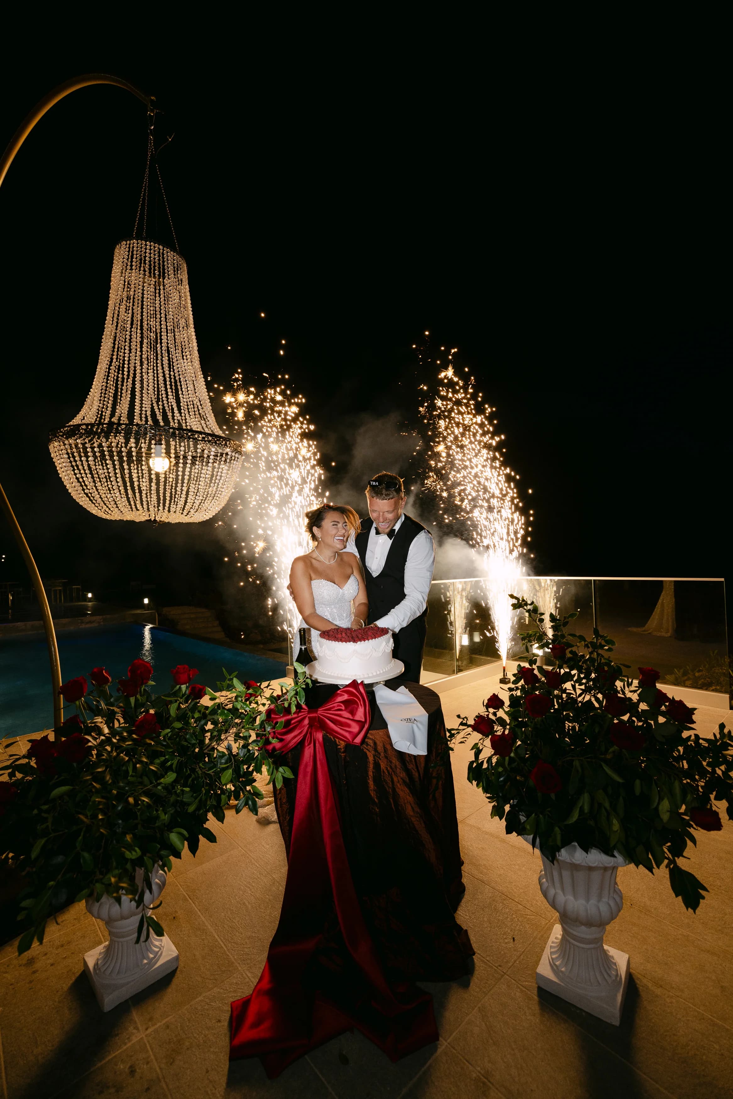 Bride and groom cutting the cake at night at OZEA with sparkler fountains behind and chandeliers framing the moment.
