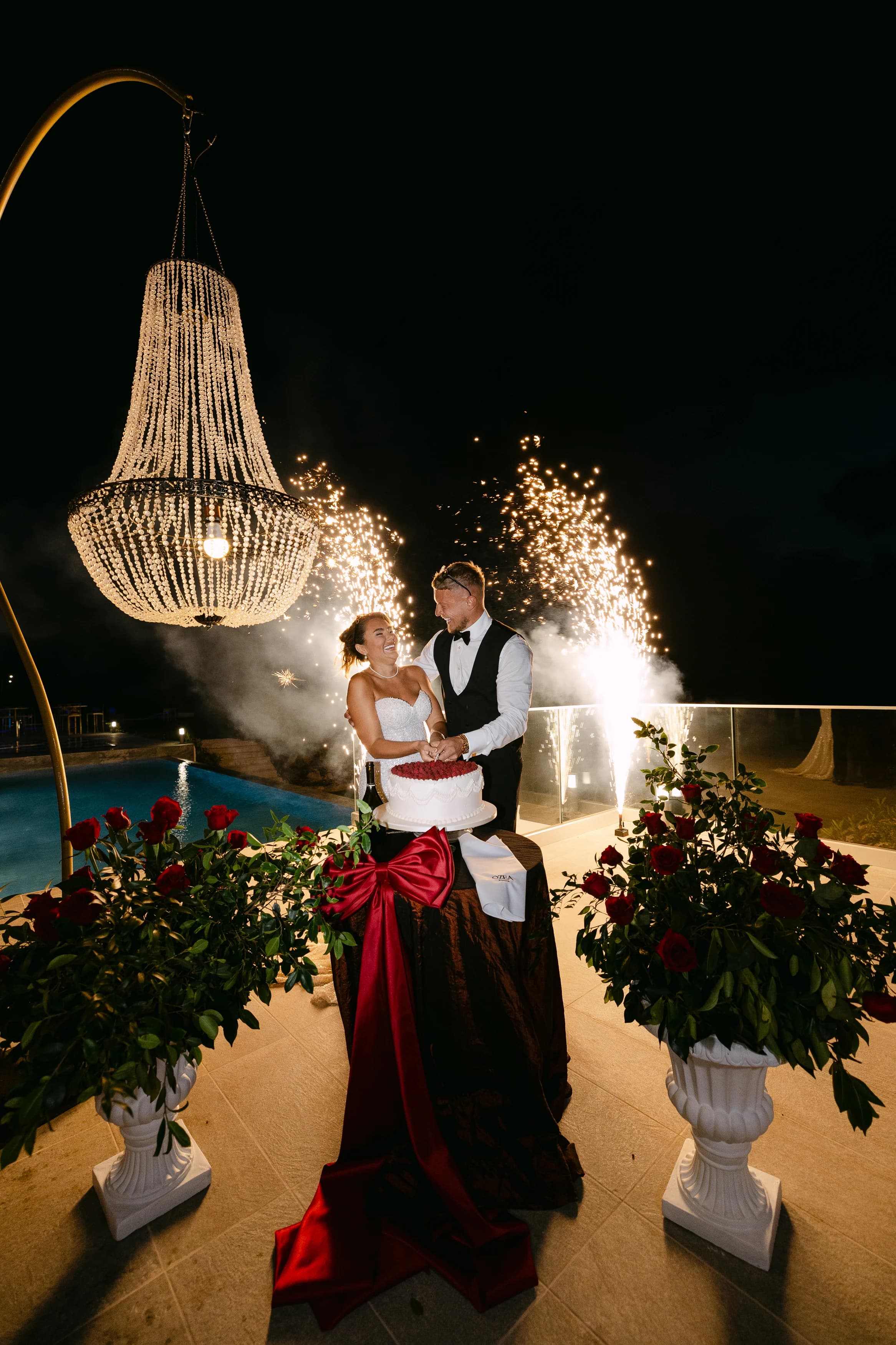 Bride and groom cutting the cake beneath a tall crystal chandelier at OZEA with sparkler fountains and red roses framing the scene.